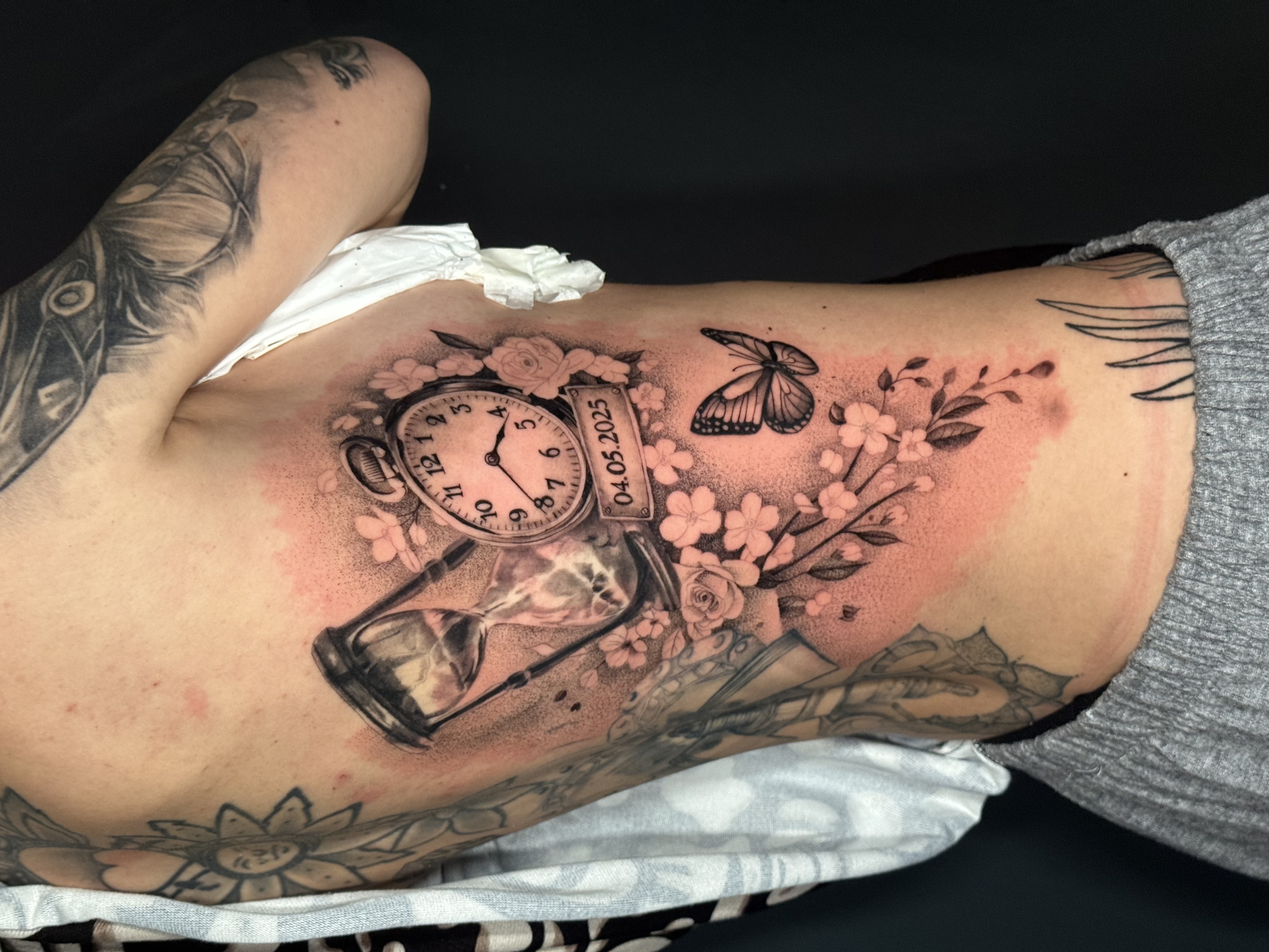 Tatuaggio black and gray con orologio e fiori sul fianco realizzato da Andrea N. Agnelli